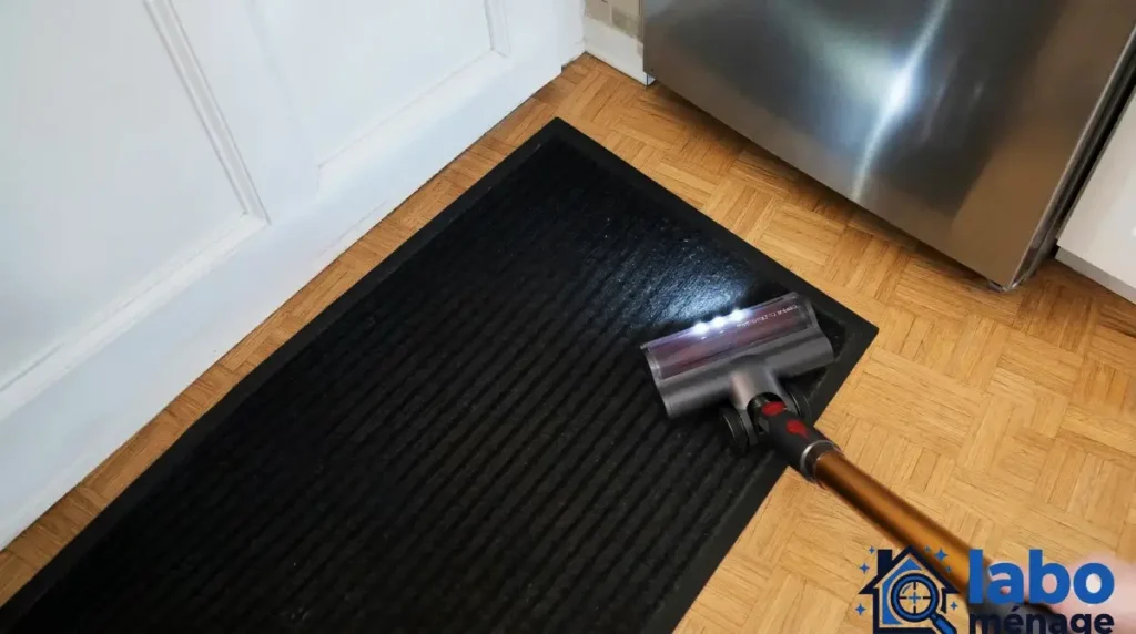 Tête de brosse posée sur un parquet, près d’un tapis noir et d’une porte.